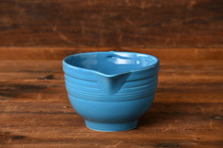 A rare and retired Peacock Batter Bowl by Fiesta Factory Direct—a blue ceramic mixing bowl with a spout—rests on a rustic wooden surface, set against a brown wood background.
