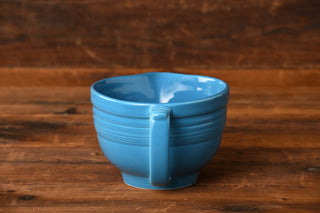 A “Peacock Batter Bowl” by Fiesta Factory Direct, in a striking blue reminiscent of the retired Peacock color from Fiesta prototypes, sits on a wooden surface with a wooden background behind it.