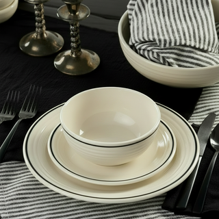 The Black Band Classic Bistro 3-Piece Place Setting by Fiesta Factory Direct, featuring a cream-colored bowl and plate with a sleek black band, is arranged on a black table alongside striped black-and-white napkins, silver cutlery, and two silver candlesticks with black candles. This elegant, minimalist table setting showcases classic bistro dinnerware made in the USA.