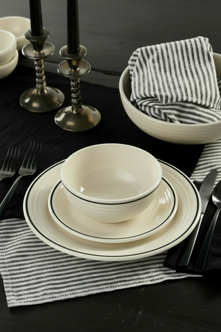 A Fiesta Black Band Classic Rim 10 1/2 Inch Dinner Plate is stacked with a white bowl and salad plate on a striped placemat beside black cutlery, with a striped napkin in the bowl and two black candles atop a black table.