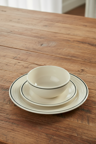 An elegant table setting showcases Fiesta dinnerware, featuring a cream-colored plate, saucer, and the Fiesta Black Band Bistro Coupe 5 1/2 Inch Cereal Bowl (22 oz) arranged on striped and black napkins and complemented by silverware. Two black candles in holders and an additional 22 oz cereal bowl with a striped napkin complete the sophisticated look on the dark tabletop.