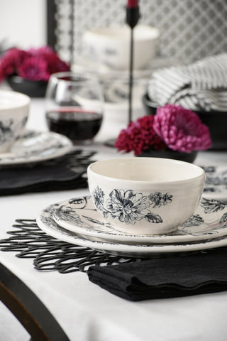 A close-up of a bistro place setting showcases the Fiesta Botanical Floral Black Classic Bistro 3-Piece Place Setting, Service For 1, featuring a beautifully patterned bowl and matching classic rim dinner plate on a black placemat with a black napkin, accented by pink flowers and a glass of red wine atop a white tablecloth.