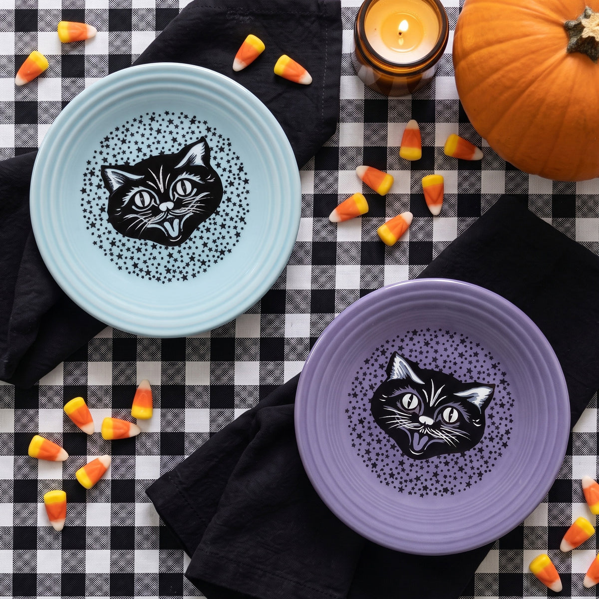 Two decorative plates with cat designs, one blue and one purple, on a checkered tablecloth with candy corn and a pumpkin.