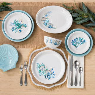 A table set with Fiesta's New Retired Bistro Coupe 3-Piece Place Setting, Service for 1 in Coastal Shores features blue and green botanical designs on white plates, complemented by blue-rimmed plates, silverware, and a shell-shaped dish on burlap placemats.