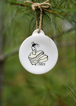 A white ceramic ornament with “2025” and a drawing of a woman in a dress is hanging from a pine branch with a twine loop. The background is blurred greenery.