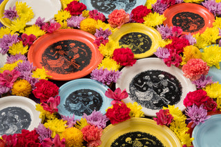 Colorful plates with black dancing skeleton illustrations in orange, yellow, white, and blue, arranged among bright flowers for a festive Day of the Dead display.