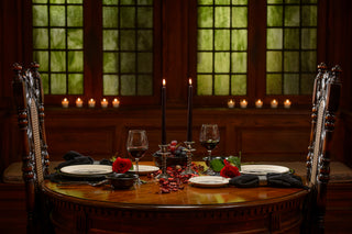 Intimate gothic-style dinner table for two, set with red roses, skeleton-themed plates, black candles, red wine glasses, and dried petals, in front of stained glass windows and candlelit ambiance.