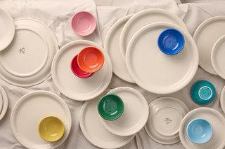 A collection of white ceramic plates of various sizes with colorful bowls—red, pink, orange, yellow, green, and blue—arranged on a soft white cloth background.