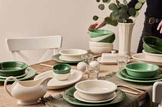 A rustic wooden dining table is set with green and white dishes, bowls, glasses, and gold cutlery. A person arranges stacked bowls near a white vase with eucalyptus branches. A white chair is by the table.