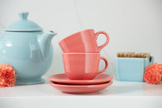 Two stacked pink ceramic cups with matching saucers placed on a white surface, next to a pale blue teapot, a box of tea bags, and orange carnations.