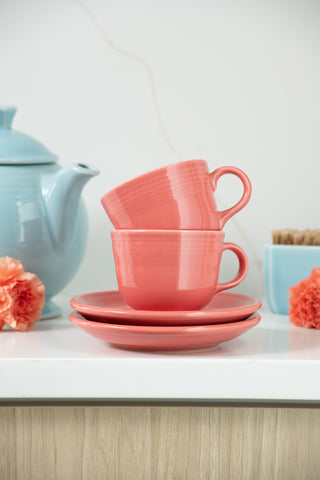 Two Fiesta (FTC) Retired Flamingo Cup & Saucer Sets, in coral, are stacked on matching plates. A blue teapot, blue bowl, and orange flowers are displayed in the background, all on a white countertop.