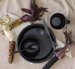 A black cup, plate, bowl, fork, knife, and spoon are arranged on a beige tablecloth with three colorful ears of corn and a folded white napkin.