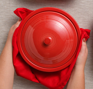 A person holds a round, bright red ceramic pot with a lid, using a matching red cloth underneath. The background is a neutral-colored fabric surface.