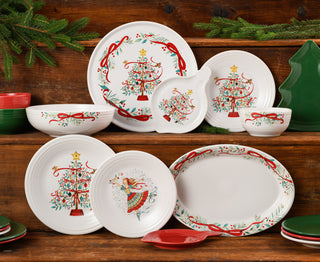 A festive set of holiday dishes with red, green, and gold designs, including Christmas trees, ribbons, and a dancing figure, displayed on wooden shelves with evergreen branches for decoration.