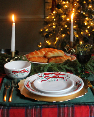 A festive holiday table features the Fiesta (FTC) Holly Berry Christmas Garland Classic Bistro Coupe 3-Piece Place Setting, gold chargers, green napkins, candles, red wine, bread rolls, evergreen branches, and a Christmas tree in the background.