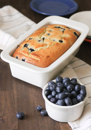 A loaf of blueberry bread sits in a white ceramic pan on a wooden table, next to a small white bowl filled with fresh blueberries. A striped kitchen towel and blue plate are in the background.