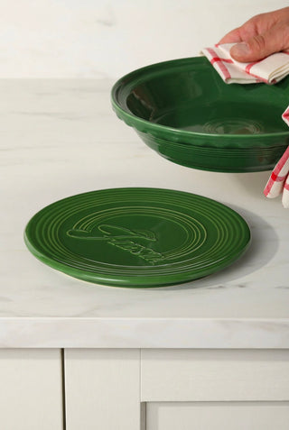 A hand holds a Fiesta Factory Direct Pie Baker and Trivet above a matching round green trivet with concentric circles on a white countertop.