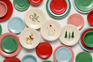 Assorted red, green, blue, and cream plates are arranged on a flat surface. Four cream plates feature festive holiday designs: a candy cane, a snowman, a reindeer, and two Christmas trees.