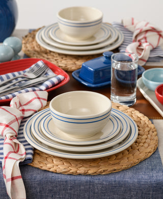 A dining table set with the Fiesta Americana Lapis Band 3-Piece Place Setting, Service for 1, featuring cream dishes with blue stripes, woven placemats, colorful trays, a blue butter dish, striped napkins, glasses of water, and a blue tablecloth.