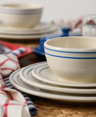 A close-up of stacked Fiesta Americana Lapis Band 3-Piece Place Setting, Service for 1—cream-colored ceramic dishes with blue stripes—on a woven placemat with red and blue checkered napkins. Proudly made in the USA.