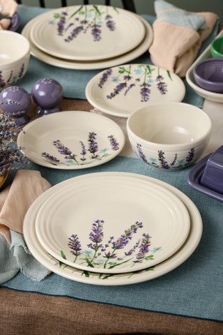 A table set with Fiesta's cream-colored dishes and bowls, featuring the Lavender Bloom Classic Rim 9 Inch Luncheon Plate, is complemented by light blue napkins, purple salt and pepper shakers, and dried lavender for an elegant touch.