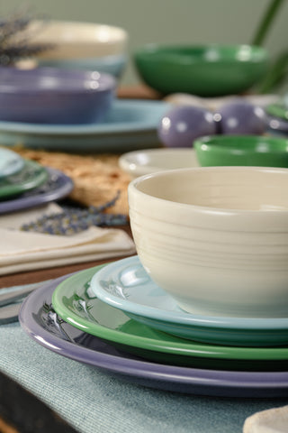 A close-up of Fiesta Factory Direct's Lavender Bloom Mixed Color Bistro Coupe 12-Piece Dinnerware Set, Service for 4, featuring stacked green, purple, and blue plates with a cream bowl, napkins, and lavender on the table; colorful dishes are in the background.