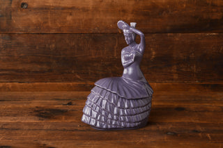 A beautiful Lavender Dancing Lady Cookie Jar by Fiesta Factory Direct features a ceramic figurine of a woman in a ruffled dress, holding a fan above her head as she stands gracefully on a wooden surface with a matching wooden background. This charming piece is perfect for any Fiesta collectible enthusiast.