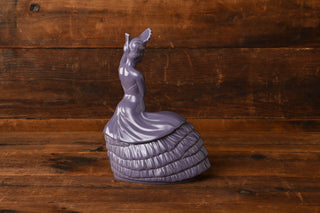 A Lavender Dancing Lady Cookie Jar by Fiesta Factory Direct, designed as a graceful purple ceramic figurine of a woman in a ruffled lavender dress and hat, sits elegantly on a wooden surface against a rustic wood background—a charming collectible for your home.
