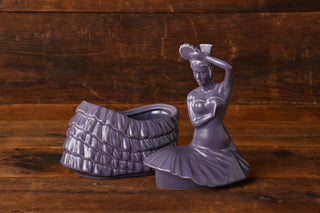 Two pieces of a Fiesta Factory Direct Lavender Dancing Lady Cookie Jar are displayed against a wooden background, with the purple ceramic figurine shaped like a woman in a dress separated into the upper body and skirt portions placed side by side.