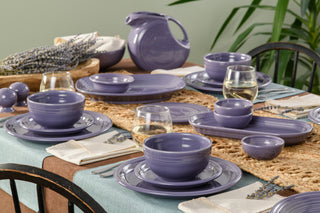 A dining table set with Fiesta’s Lavender Bistro Coupe 3-Piece Place Setting, Service for 1, featuring modern dishes in purple hues, clear white wine glasses, linen napkins, a woven placemat, sprigs of lavender, and a plant in the background.