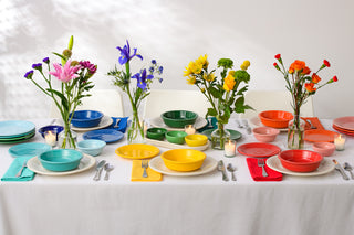 A table set with colorful plates, bowls, and napkins, each place setting in a different color. Small vases with vibrant flowers and candles are arranged along the center of the table on a white tablecloth.