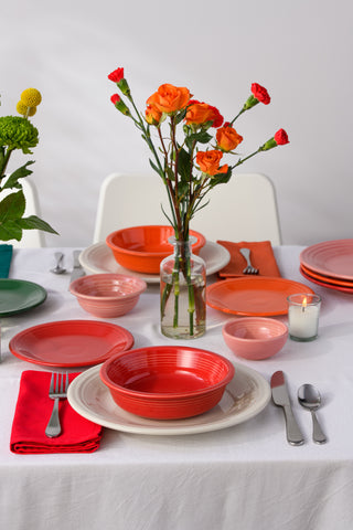 A table set with colorful plates and bowls in shades of red, pink, and green, silver cutlery, a red napkin, a lit candle, and vases with orange and yellow flowers on a white tablecloth.