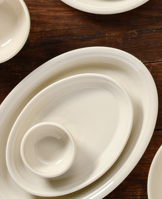 Several cream-colored ceramic plates and bowls of different sizes are arranged on a wooden surface, overlapping each other. The dishes have a simple, smooth design with subtle concentric rings.