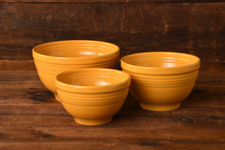 Three yellow ceramic nesting bowls of varying sizes are arranged on a rustic wooden surface, with a wooden wall in the background—perfectly showcasing the Marigold 3pc Baking Bowl Set by Fiesta Factory Direct, a beautiful tribute to Fiesta’s 75th Anniversary.
