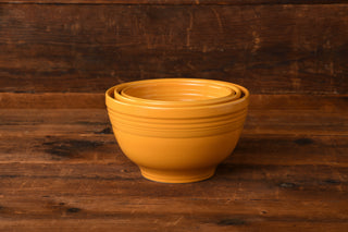 Three yellow ceramic bowls from the Fiesta Factory Direct Marigold 3pc Baking Bowl Set, nested inside each other, are displayed on a rustic wooden surface with a wooden background—a timeless tribute to this collection’s 75th Anniversary.