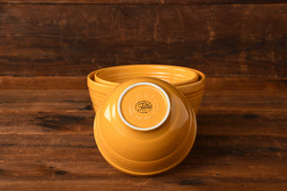 Three nesting yellow bowls from the Marigold 3pc Baking Bowl Set by Fiesta Factory Direct rest on a wooden surface. The base of the top bowl faces forward, displaying the Fiesta Made in USA brand marking. This charming set is presented against a rustic wooden wall.