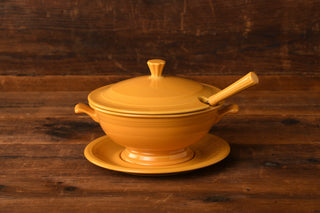 A Marigold 75th Anniversary Soup Tureen by Fiesta Factory Direct, complete with a lid, matching ladle, and coordinating plate in a bold marigold yellow, is displayed on a wooden surface against a rustic wood background. This limited edition piece from Fiesta Factory Direct’s coveted collection adds timeless charm and collectible appeal to any table setting.