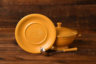 A yellow plate embossed with Fiesta 75th Anniversary, a coordinating covered dish from the Fiesta Factory Direct museum collection, and a ladle rest on a rustic wooden surface highlight the charm of the Marigold 75th Anniversary Soup Tureen by Fiesta Factory Direct.