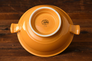 A Marigold 75th Anniversary Soup Tureen by Fiesta Factory Direct is positioned upside down on a wooden surface, displaying the Fiesta brand logo and “USA” stamped on its base, with two handles in view.