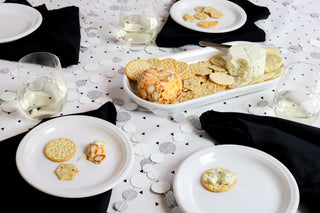 A table set with white plates and glasses of white wine, featuring a platter of assorted crackers and two cheese balls. Black napkins and a white tablecloth with silver dot patterns complete the setting.