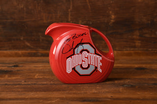 A Fiesta Factory Direct Ohio State Fiesta Large Disk Pitcher, signed by Urban Meyer in black marker and featuring the Ohio State logo, is displayed on a wooden surface with a wooden background—an impressive collectible from the Upper Ohio Valley Museum.