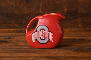 A Fiesta Factory Direct Ohio State Fiesta Large Disk Pitcher, signed by Urban Meyer, features a red ceramic design with the Ohio State logo and text and is displayed on a wooden surface against a wooden background. This distinctive collectible is a highlight of the Upper Ohio Valley Museum’s collection.