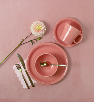 A pink dinnerware set with plate, bowl, saucer, and cup, arranged with a gold fork, knife, and spoon on a pink surface. A white flower and scalloped napkin complete the setting.