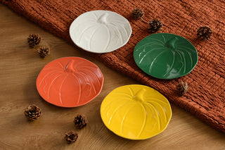 Four pumpkin-shaped plates in white, green, orange, and yellow are arranged on a wooden surface with pinecones, next to a textured orange cloth.