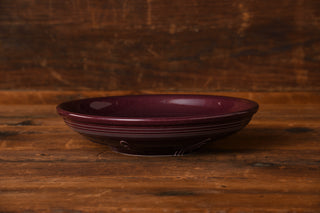 A Raspberry Presentation Bowl by Fiesta Factory Direct, featuring an exclusive dark purple glaze and a shallow ceramic design, is displayed on a rustic wooden surface with a brown wood background.