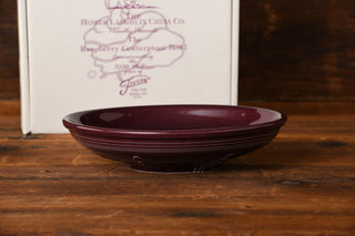 A deep, round Raspberry Presentation Bowl from Fiesta Factory Direct—its dark purple hue echoing the exclusive Raspberry glaze beloved by collectors—rests on a wooden surface, while a white box adorned with purple printed text and design stands in the background.