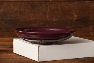 A Raspberry Presentation Bowl by Fiesta Factory Direct, featuring an exclusive Raspberry glaze in a deep purple hue, rests atop a closed white box on a wooden surface with a rustic wooden background.
