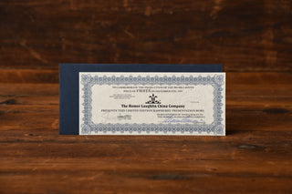 A blue and white certificate from Fiesta Factory Direct rests on a wooden surface with a dark blue envelope partially visible behind it, commemorating the exclusive Raspberry glaze featured on this limited-edition Raspberry Presentation Bowl collectible.
