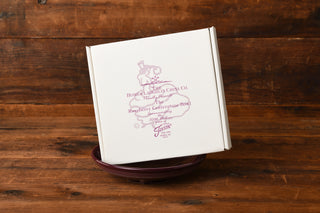 A white box labeled Fiesta Factory Direct Raspberry Presentation Bowl, featuring the exclusive Raspberry glaze, sits tilted on top of a maroon bowl against a wooden background.