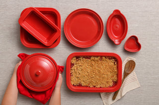 Collection of bright red ceramic bakeware including casserole dishes, plates, and heart-shaped bowls, with a baked dessert in a rectangular dish and wooden spoon nearby.